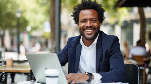 A smiling person seated at a café table with a laptop, exhibiting a relaxed and confident demeanor.