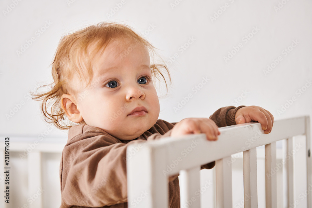 portrait of adorable child standing and looking away in crib in nursery room, happy babyhood