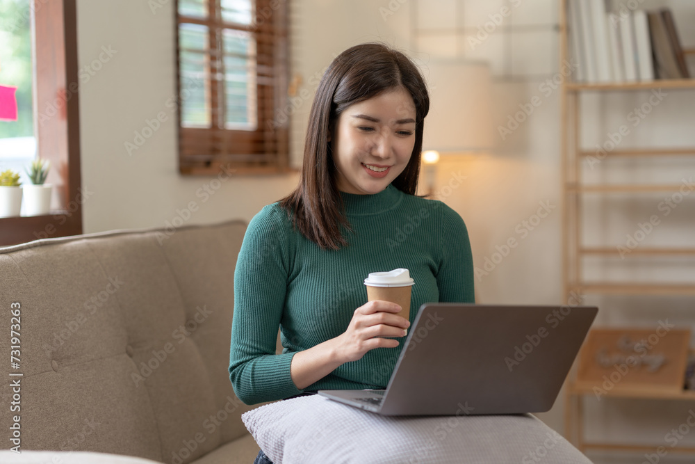 A young professional asian woman smiling while working on a laptop, holding a coffee cup in a cozy home office. A young professional asian woman smiling while working on a laptop, holding a coffee cup in a cozy home office.