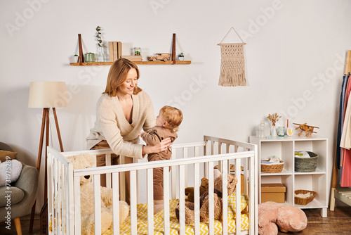 joyful young mother holding cute toddler son near crib in nursery room, blissful motherhood