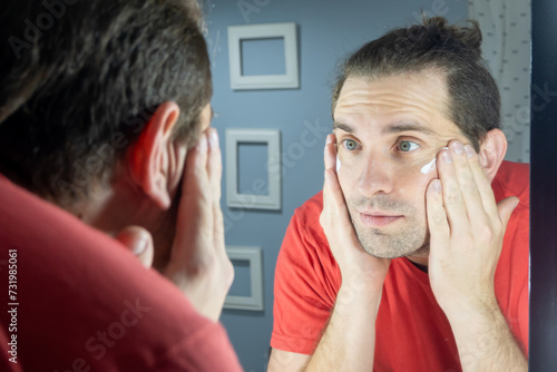 Skin care man. Man applies cream to his face in front of the mirror. Daily skin care for a man. Natural photo at home.