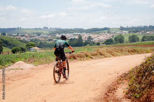 Ciclista de montanha pedalando por estrada rural, cidade desfocada ao fundo, e céu azul.