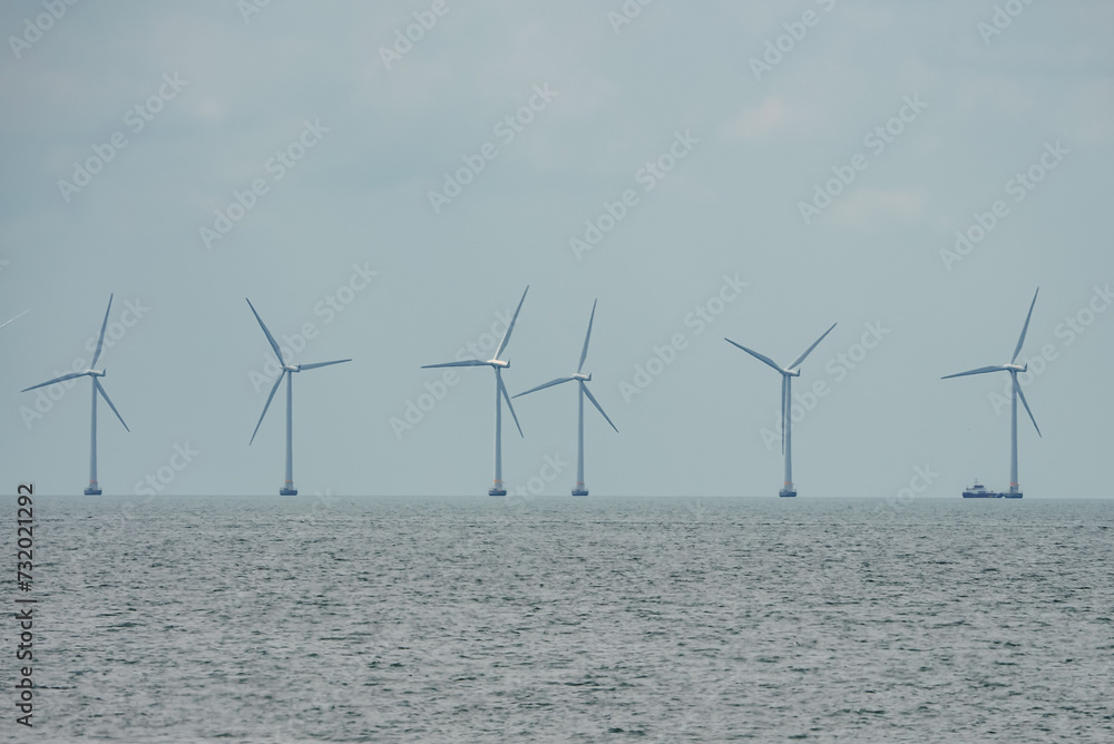 Large wind turbines rotate in the calm sea under an overcast sky, lined up towards the horizon in the Oresund Strait between Copenhagen and Malmo, with a boat near the last turbine for scale.