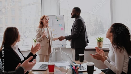 Boss shaking hand of young shy woman congratulating successful employee with promotion, hiring intern, appreciating for good work result, rewarding while business team applauding supporting colleague