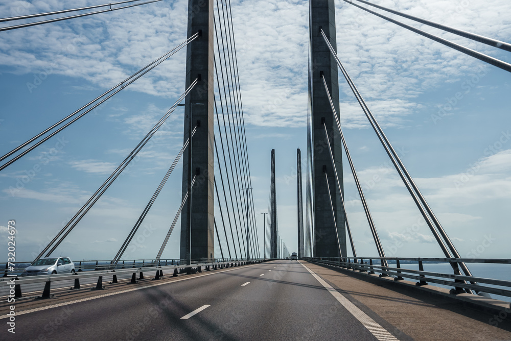 Obraz premium A clearskied modern suspension bridge with twin towers and cables, calm seas on either side. An empty lane, white car seen, possibly the Oresund Bridge linking Copenhagen to Malmo.