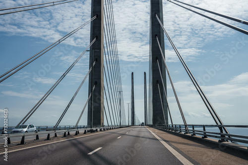 A clearskied modern suspension bridge with twin towers and cables, calm seas on either side. An empty lane, white car seen, possibly the Oresund Bridge linking Copenhagen to Malmo. © Aerial Film Studio