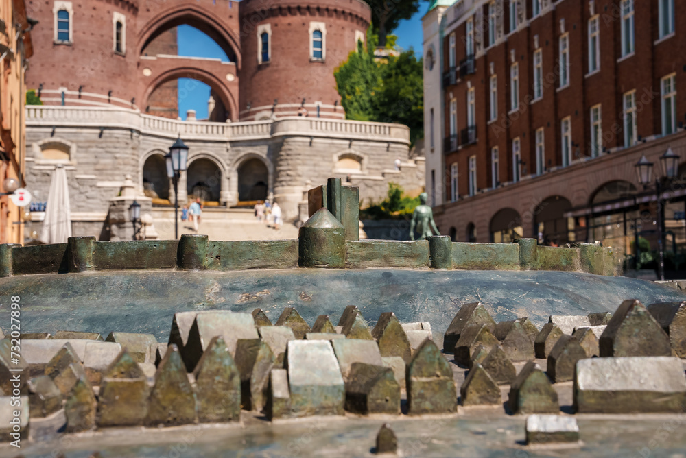 Fototapeta premium An urban scene with a dormant fountain in the foreground and a blurred background featuring a redbrick building with arches, a clear blue sky, and hints of nearby cafes in Helsingborg.