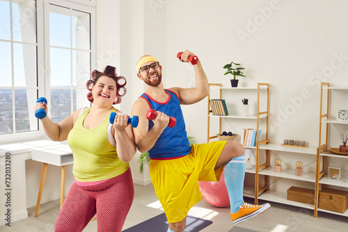 Funny overweight happy fat woman with bearded man in bright sportswear looking at the camera and doing fit exercises with dumbbells at home together. Concept of couple family sport and fitness.