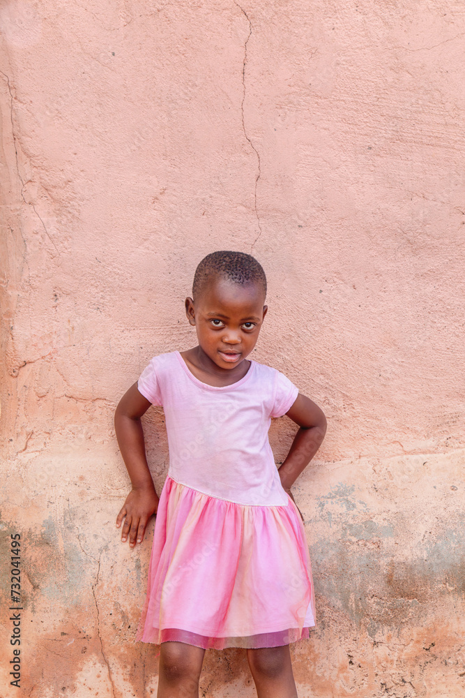 village, african girl dressed in pink in front of the house. holding onto the wall village, african girl dressed in pink in front of the house. holding onto the wall