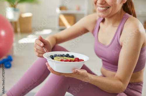 Cropped portrait of a young sporty cheerful slim woman wearing sportswear eating healthy food after working out at home sitting on the floor. Healthy nutrition and lifestyle concept.