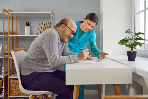Smiling young female nurse, doctor or caregiver consulting cheerful senior man client. Mature man signing insurance contract at the desk at home. Elderly healthcare and help concept.