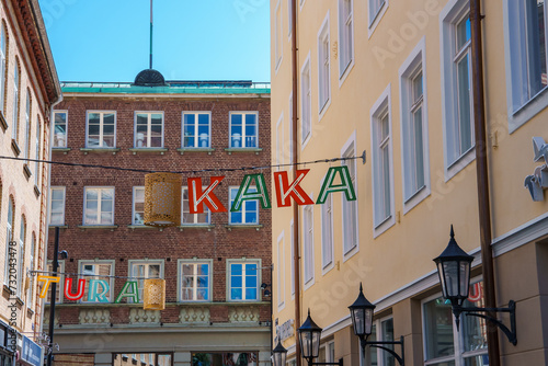Europeanstyle buildings line a serene street under a clear sky, with colorful letters spelling KAKA and TURA strung overhead, and classic lanterns adding charm, possibly in Helsingborg. photo