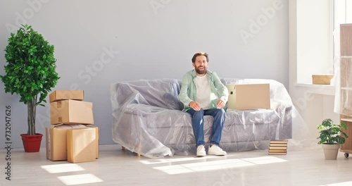 Young man buys and moves to new house or apartment. Happy man with cardboard box enters living room, puts box on sofa covered with protective film, sits down, puts hands behind head and looks around photo