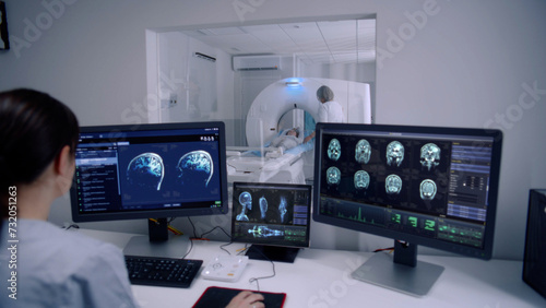 Female radiologist sits in control room and watches at monitors with displayed brain scans results. Professional doctor controls MRI or CT or PET scan with patient undergoing procedure at background.