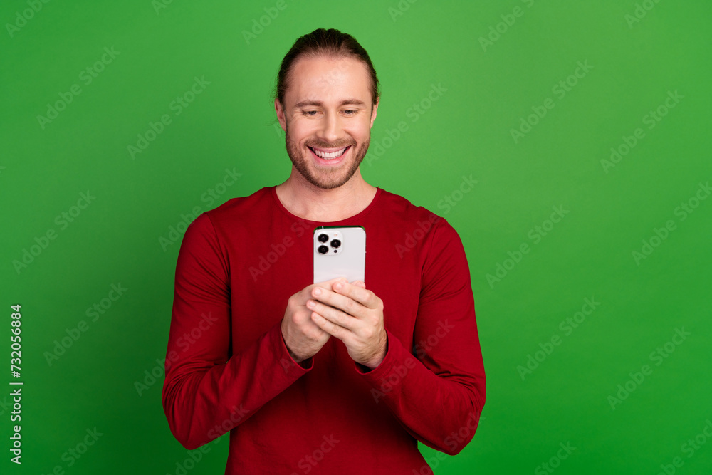 Portrait of smart clever guy with long hair wear red shirt choose product in eshop on smartphone isolated on green color background Portrait of smart clever guy with long hair wear red shirt choose product in eshop on smartphone isolated on green color background