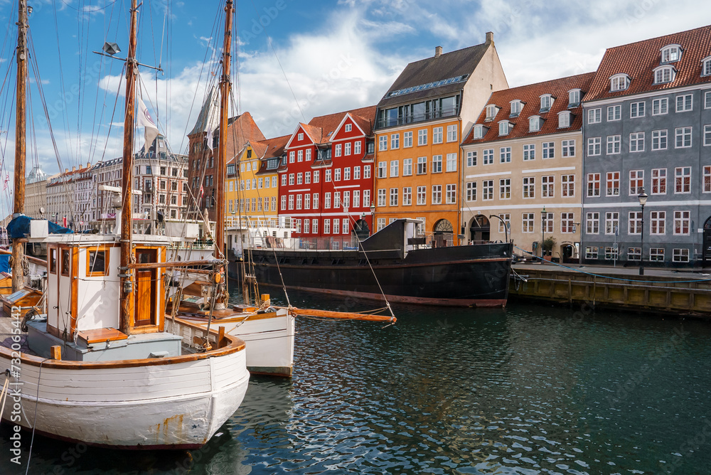 Fototapeta premium Copenhagen's calm harbor, with traditional boats and a white wooden vessel, set against Nyhavn's colorful facades under a semicloudy sky, radiates a lively cultural allure.