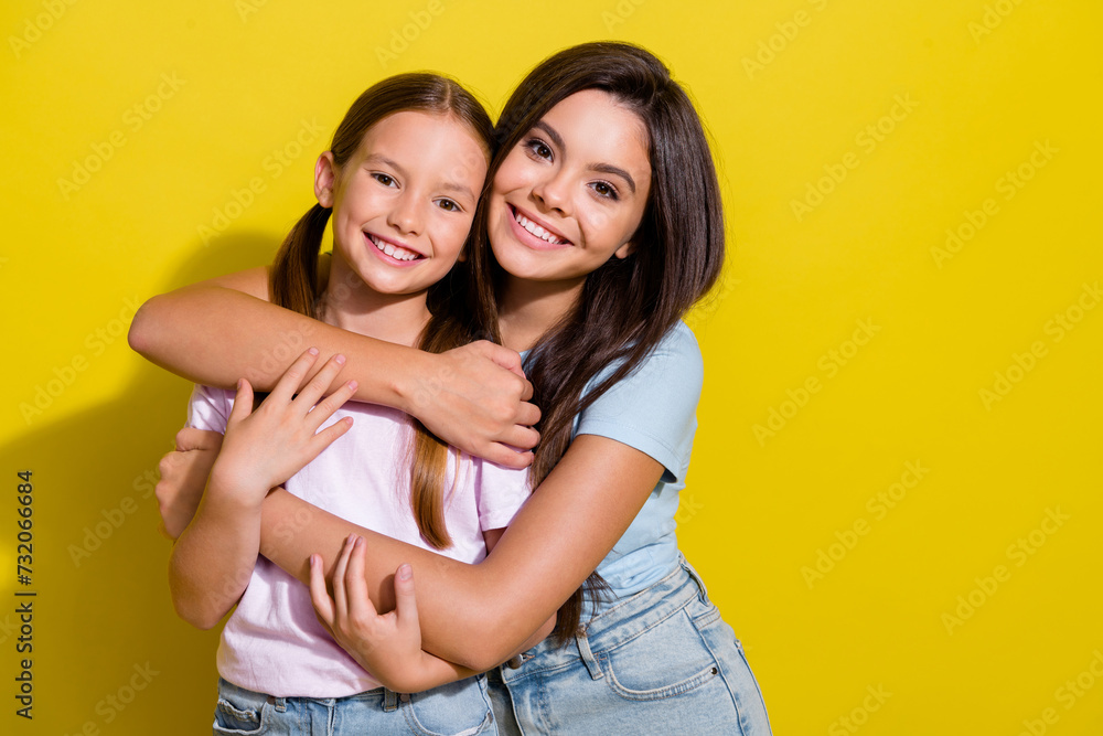 Photo of pretty cheerful little sisters wear t-shirts cuddling smiling empty space isolated yellow color background Photo of pretty cheerful little sisters wear t-shirts cuddling smiling empty space isolated yellow color background