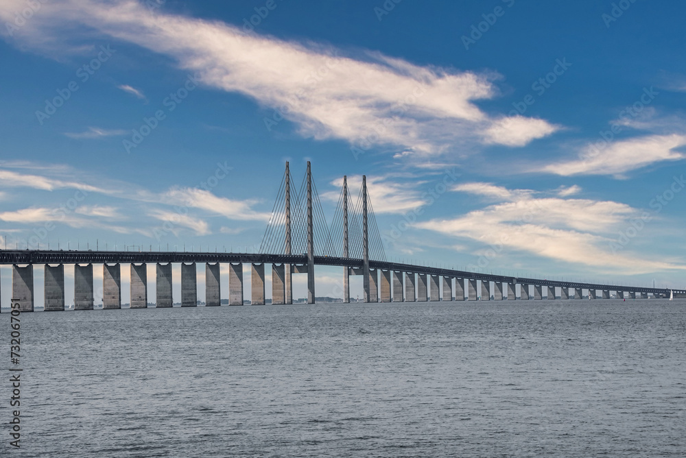 The Oresund Bridge, connecting Copenhagen, Denmark, with Malmo, Sweden, arches over calm waters, its high pylons and cables creating a striking silhouette against a clouddotted sky.