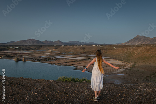 Girl in white dress stands on a cliff and looks on the Janubio flats