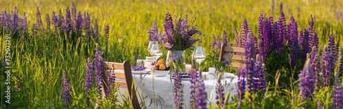 Elegant gorgeous wedding table decor or romantic dinner arrangement outdoors in blooming field. Purple lupine flowers, candles, fruits and wine, wooden furniture. Sunset, summer, golden hour banner