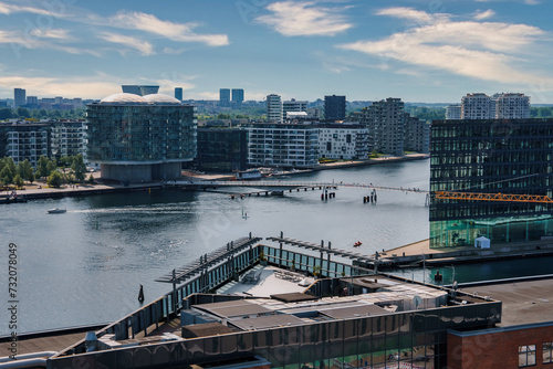 Panoramic daytime view of Copenhagen s modern waterfront with clear skies  reflecting water  boats  and locals on a pier. Contemporary architecture and ongoing construction highlight urban growth.