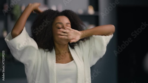 Morning African American beautiful woman come in bathroom at home looking at mirror reflection stretching after wake up look at herself ethnic girl lady female stretch yawn pampering touch face hair photo