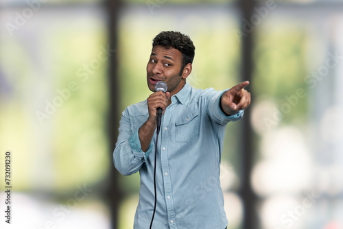 Funny indian man entertainer talking into microphone and pointing with index finger. Blur interior background.