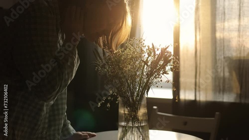 Young cheerful woman is enjoying smell of beautiful bouquet of wild flowers in glass vase on table in sunlight from window at home kitchen. Gift and celebration valentines day, birthday, Women's day.