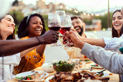 Young group of people enjoying dinner reunion in the evening at rooftop home. Millennial diverse friends social gathering reunited on terrace table, having fun celebrating barbecue party at night. photo