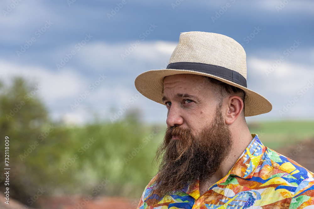 Young bearded man in fedora hat standing smiling happily. Close-up portrait of a bearded young man wearing casual clothes looking at the camera with a cheerful face in a grassy field.