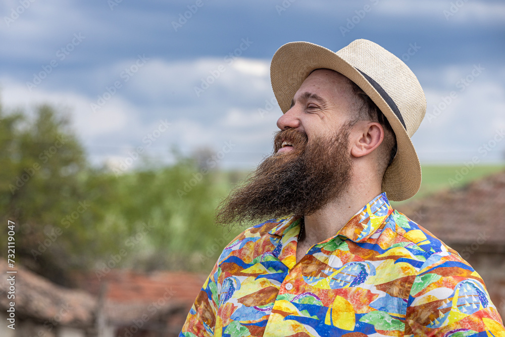 Young bearded man in fedora hat standing smiling happily. Close-up portrait of a bearded young man wearing casual clothes looking at the camera with a cheerful face in a grassy field.