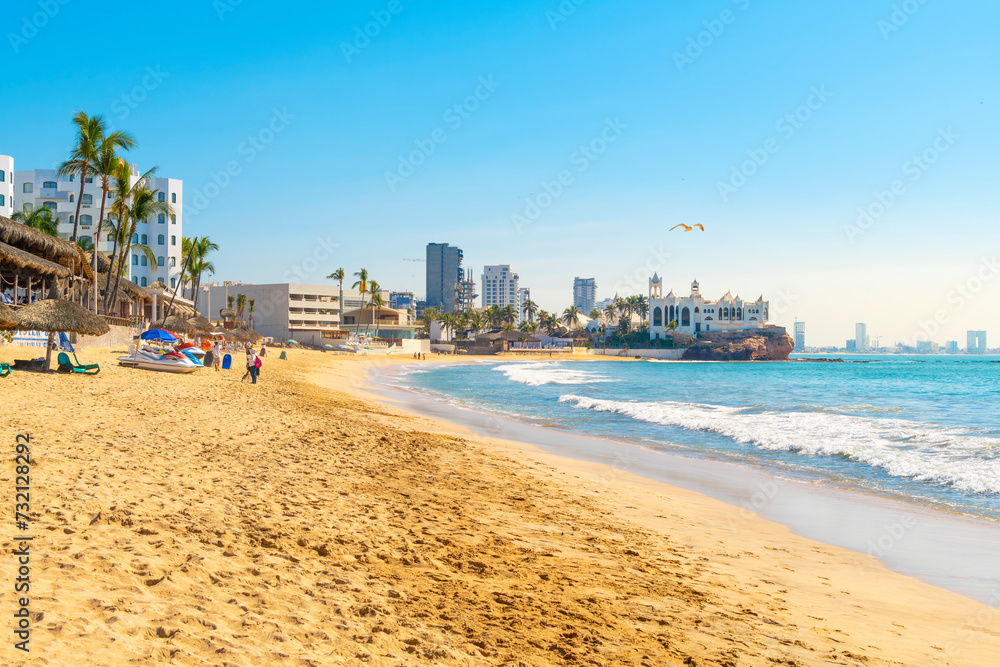 The wide sandy Playa Gaviota beach full of resorts and restaurants, with Valentinos Disco and the skyline of Mazatlan, Mexico behind, on a sunny day along the Golden Zone.