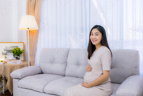 Smiling beautiful pregnancy woman sitting on sofa by the window of her home, showcasing beauty and a positive lifestyle.Asian pregnant female.White background with copy space.Hand on tummy or belly.