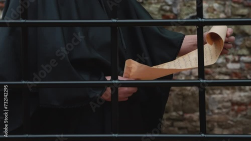 A monk in a black cassock unrolls a scroll and reads behind a grille against a stone wall. photo