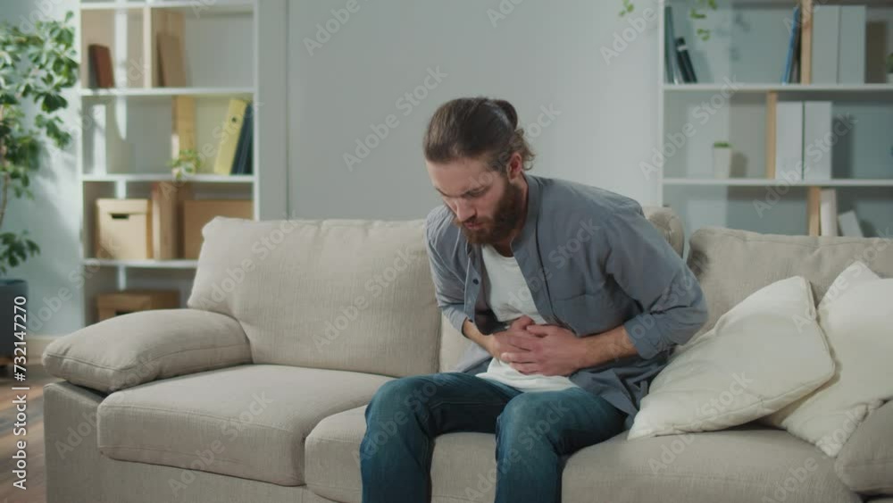 Man sitting on the couch suddenly felt pain in his stomach. Man trying to relieve pain by massaging his stomach Man sitting on the couch suddenly felt pain in his stomach. Man trying to relieve pain by massaging his stomach