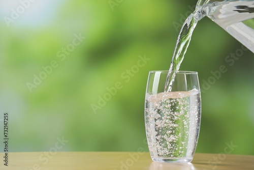 Pouring fresh water from jug into glass at wooden table against blurred green background, closeup. Space for text