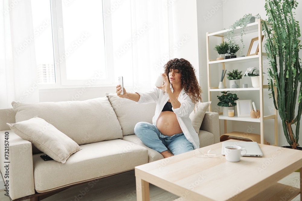 Pregnant woman blogger with a jar of pills and vitamins sits on the couch at home and takes pictures of herself on the phone, maintaining pregnancy and replenishing vitamins and minerals Pregnant woman blogger with a jar of pills and vitamins sits on the couch at home and takes pictures of herself on the phone, maintaining pregnancy and replenishing vitamins and minerals
