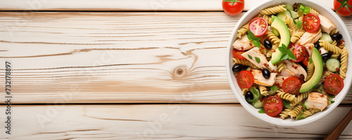 From above, a white bowl holds a delicious Chicken Pasta Salad adorned with avocado, tomato, and dressed with olive oil and vinegar on a white wood table. A visually appealing image, ideal for creativ photo