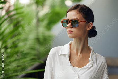 Smiling woman enjoying a tropical vacation on a balcony She is dressed in trendy fashion, wearing sunglasses, and basking in the sunlight With a refreshing drink in hand, she stands on the terrace of