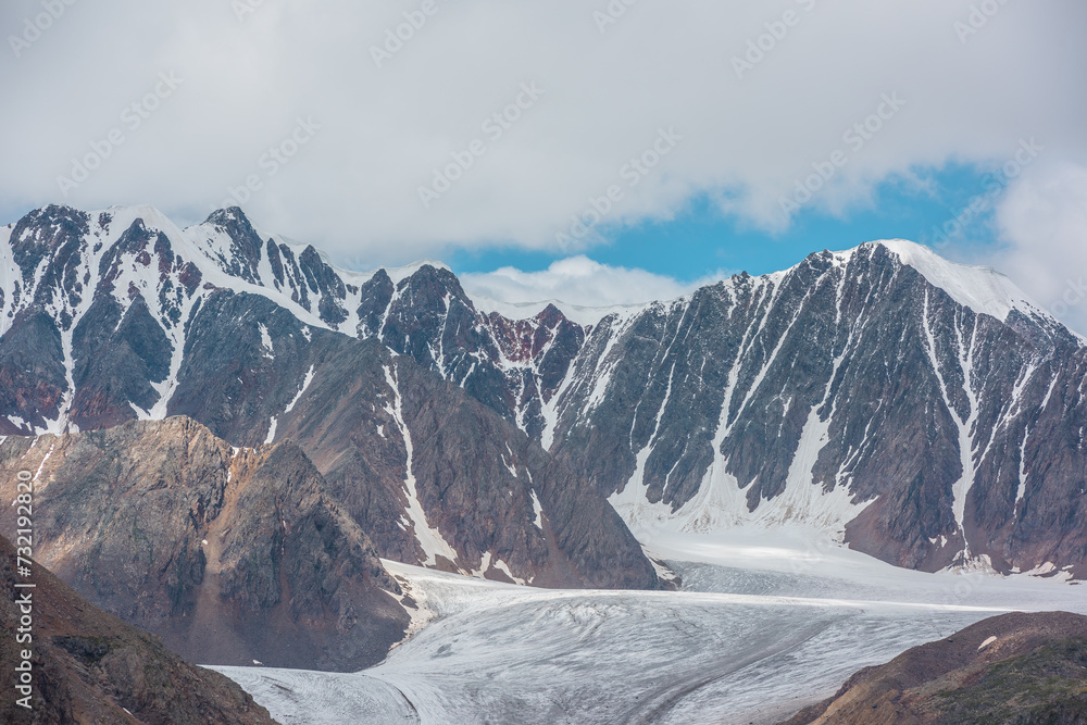 Dramatic view to beautiful glacier tongue among rocky ridges and sharp snow-capped mountain range in sunlight under clouds in blue sky. Ice and sheer crags close up. Snow cap on high rock mountain.