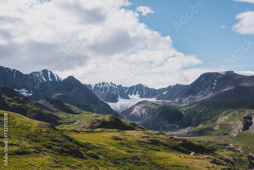 Dramatic scenery in alpine valley with creek among green hills and rocks with view to rocky pointy peak, large snow-capped peaked top, mountain range and big glacier tongue under clouds in blue sky.