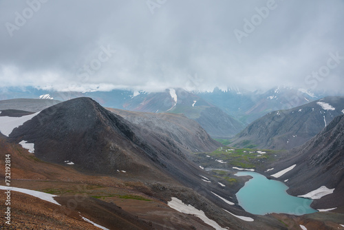 Most beautiful alpine lake in stony green valley and rocky black hill silhouette in low clouds. Mountain range in gray sky in far away. Large snowy mountains in light transparent mist in rainy weather