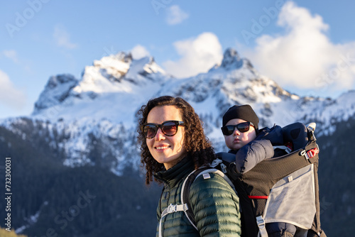 Happy Mother with Baby Boy in a Hiking Backpack Carrier with Mountains in the Background. Sunny Winter Sunset. Squamish, BC Canada.