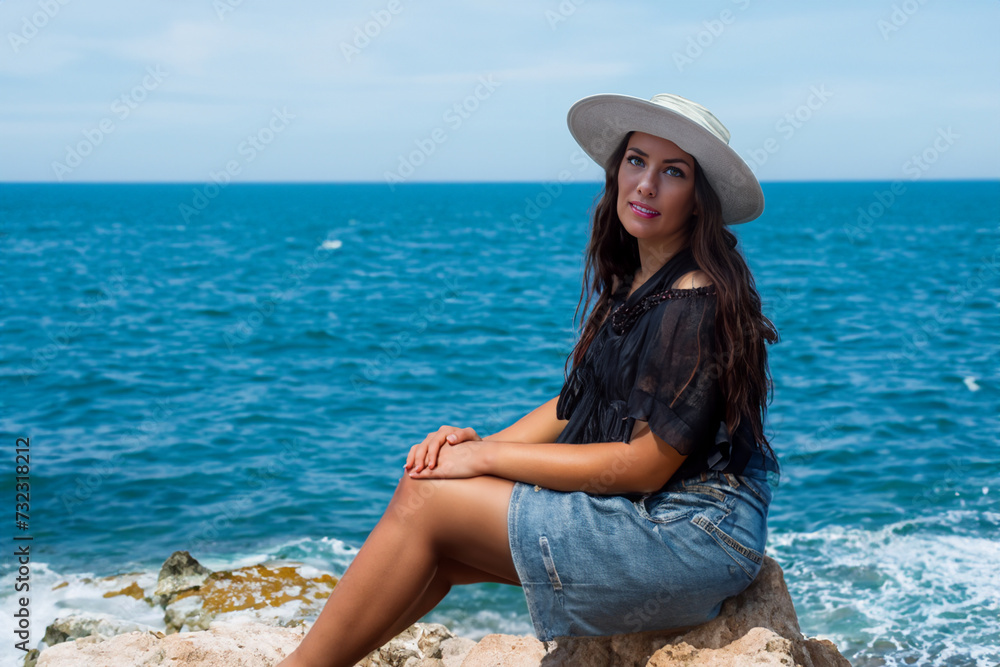 woman on vacation. happy woman with dark long hair, wearing a black T-shirt and denim skirt, sitting on a stone near the sea with a hat on her head, vacation concept