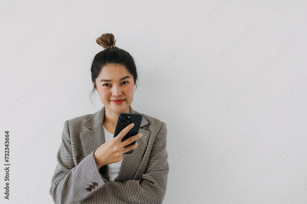 Asian Thai woman happy using mobile phone, smiling at camera while holding smartphone, wear blazer suit smart look, standing isolated over white background wall. Asian Thai woman happy using mobile phone, smiling at camera while holding smartphone, wear blazer suit smart look, standing isolated over white background wall.