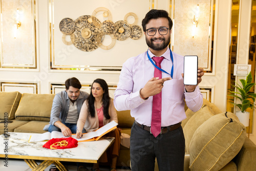 Happy looking indian Bank Agent showing mobile, smart phone. couple doing documentation process for gold loan in background with gold jewelery. Finance and Loan Concept.