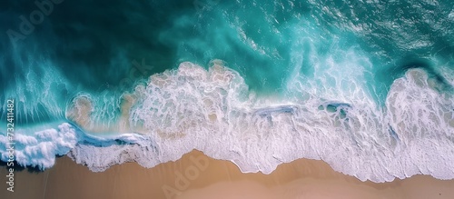 An artistic display of electric blue waves crashing on a sandy beach, witnessed from an aerial view. The sky, adorned with cumulus clouds, adds to the beauty of this meteorological phenomenon.