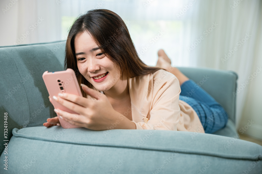 Smiling woman relaxed social online communication on smartphone, Happy Asian female using smart mobile phone lying on sofa in living room room at house, technology gadget online application Smiling woman relaxed social online communication on smartphone, Happy Asian female using smart mobile phone lying on sofa in living room room at house, technology gadget online application