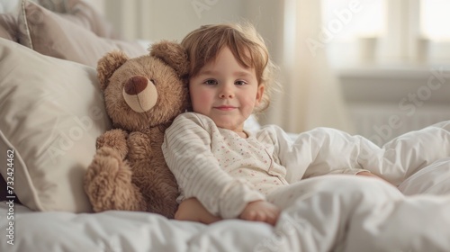 A young child with a joyful expression lying on a bed with a large brown teddy bear surrounded by soft pillows and blankets in a warm and cozy setting.
