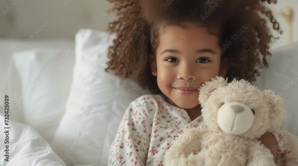 A young girl with curly hair wearing a floral nightgown smiling and holding a white teddy bear sitting on a white bed with white pillows. A young girl with curly hair wearing a floral nightgown smiling and holding a white teddy bear sitting on a white bed with white pillows.